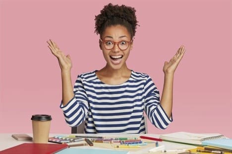 Amazed black female illustrator raises hands in eureka gesture, wears striped clothes